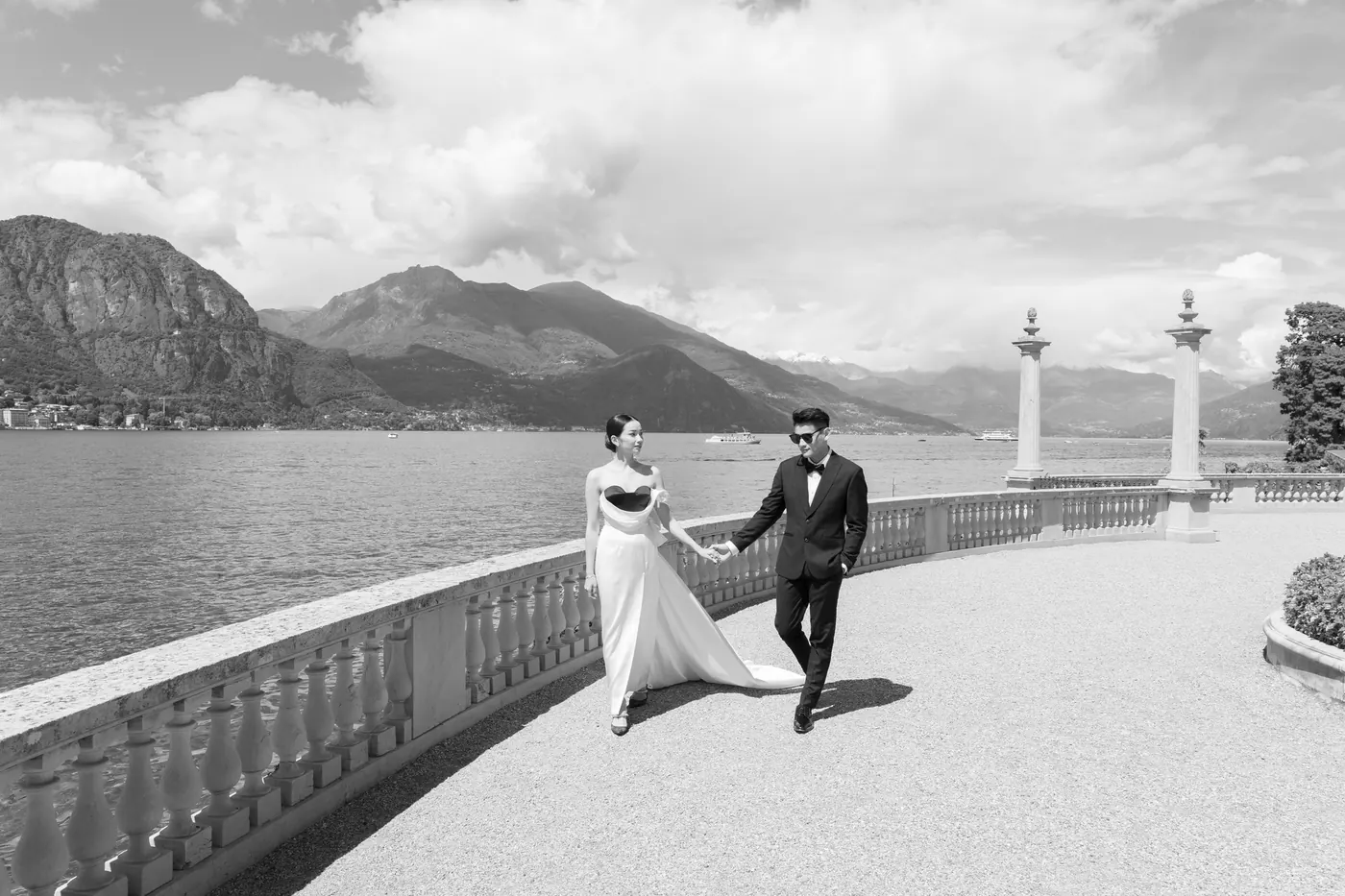 Matthew and Karin walking hand in hand along a lakeside balustrade, mountains behind, in black and white